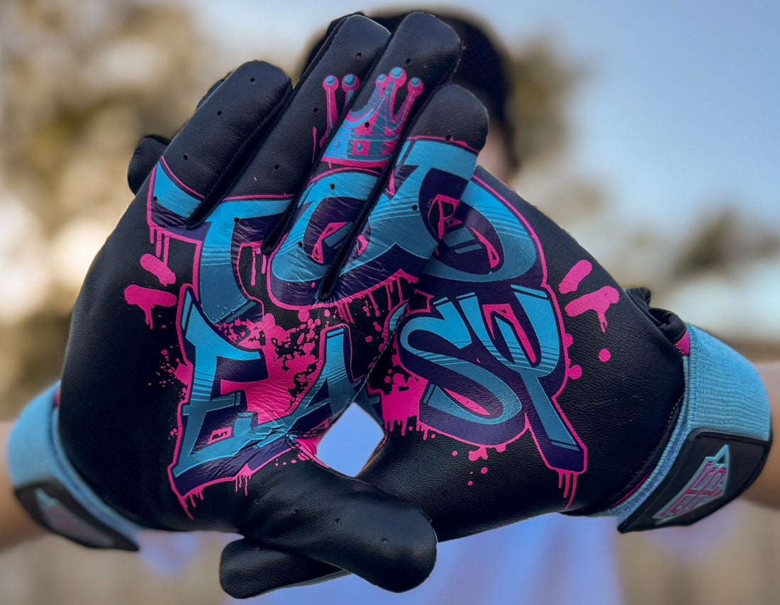 A pair of Candy Crushers baseball batting gloves with a 'Comic Hands' design in blue and pink colors, displayed in a neon-lit urban environment.